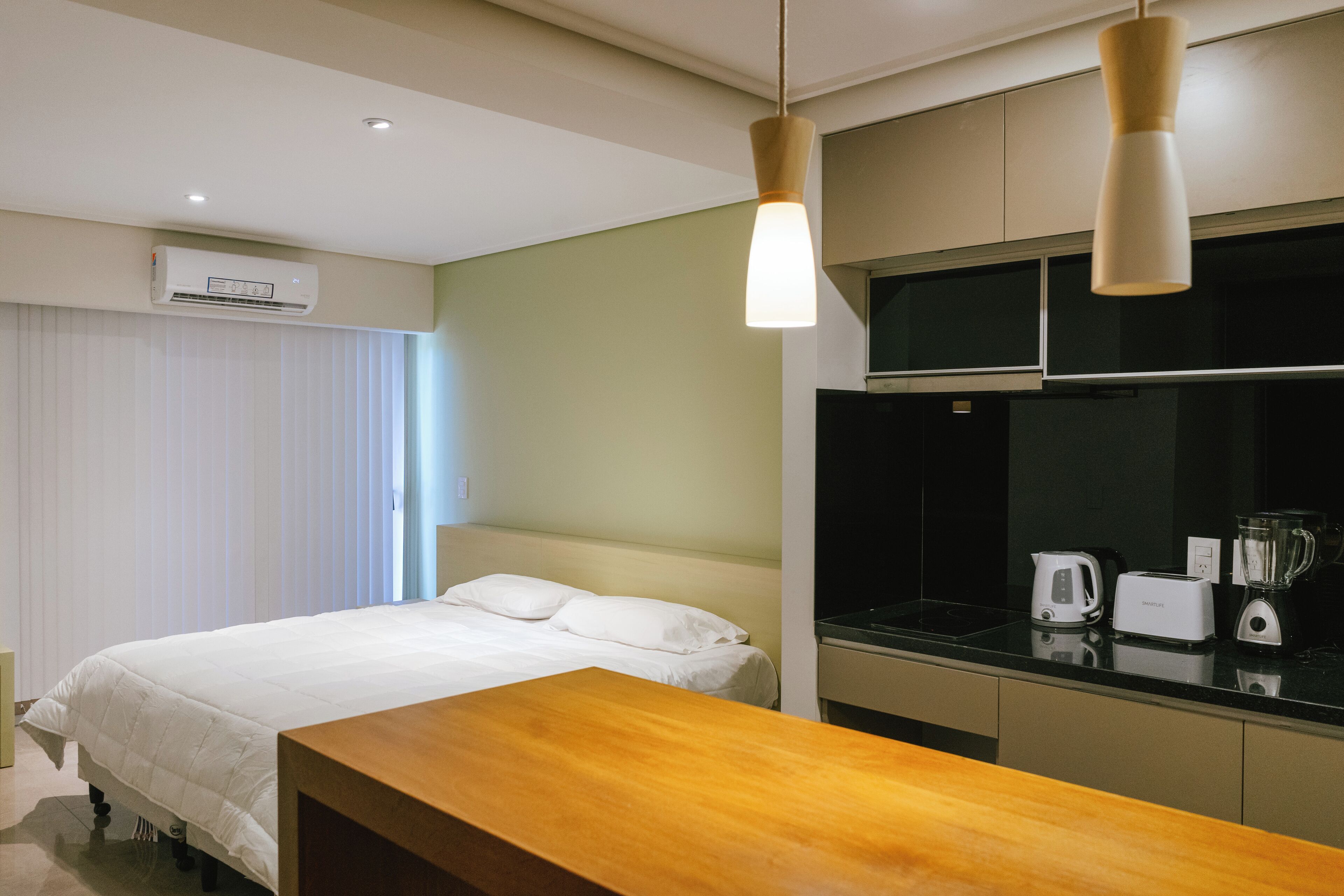 A hotel room with a bed, a wooden bench, a kitchenette with a kettle, toaster, and blender, and an air conditioning unit.