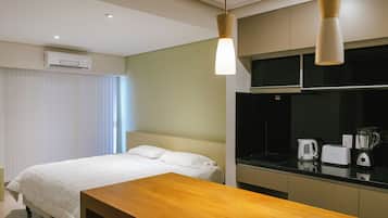 A hotel room with a bed, a wooden bench, a kitchenette with a kettle, toaster, and blender, and an air conditioning unit.