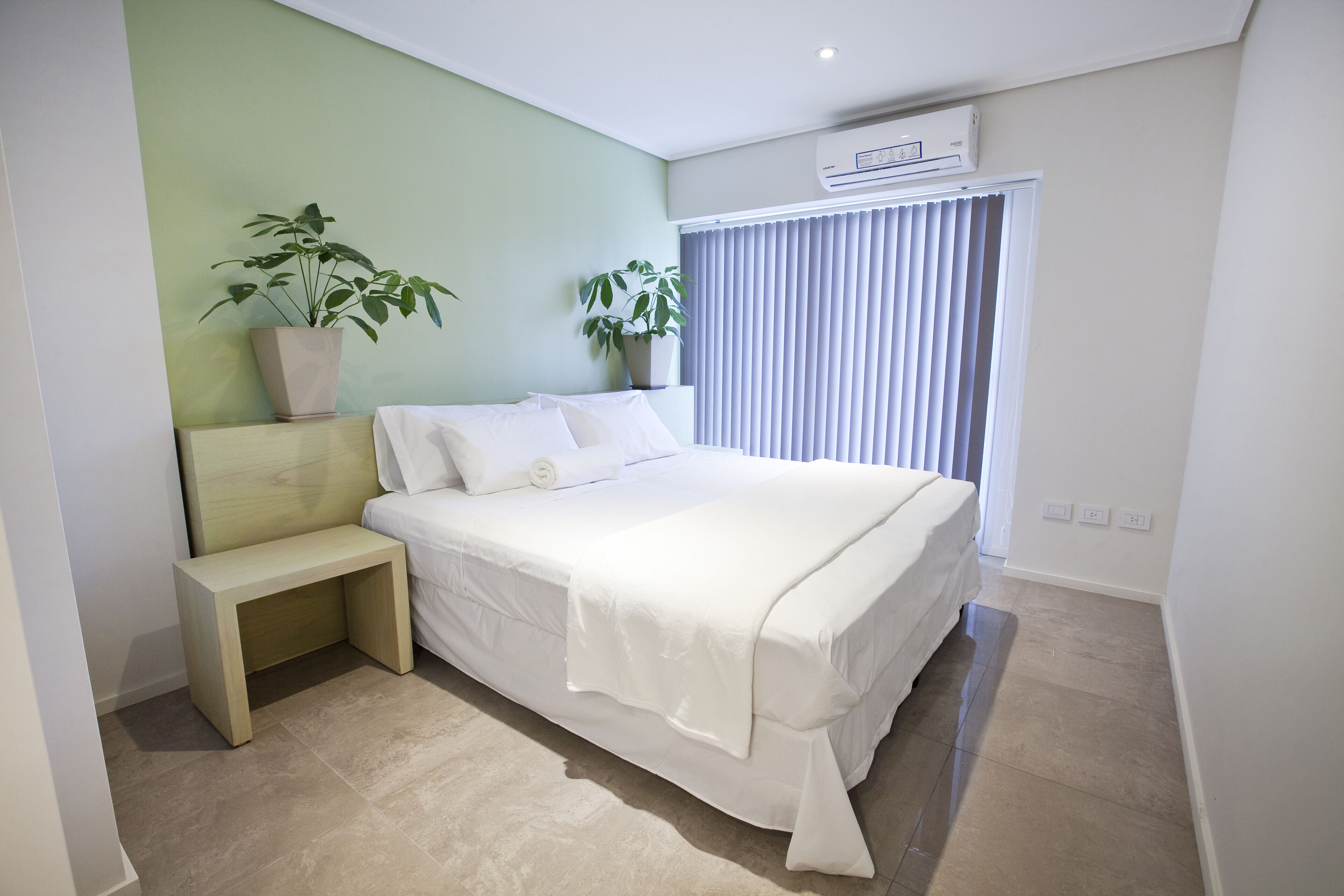 Un dormitorio con cama, mesitas de noche, dos plantas en macetas, una ventana con persianas y un equipo de aire acondicionado.