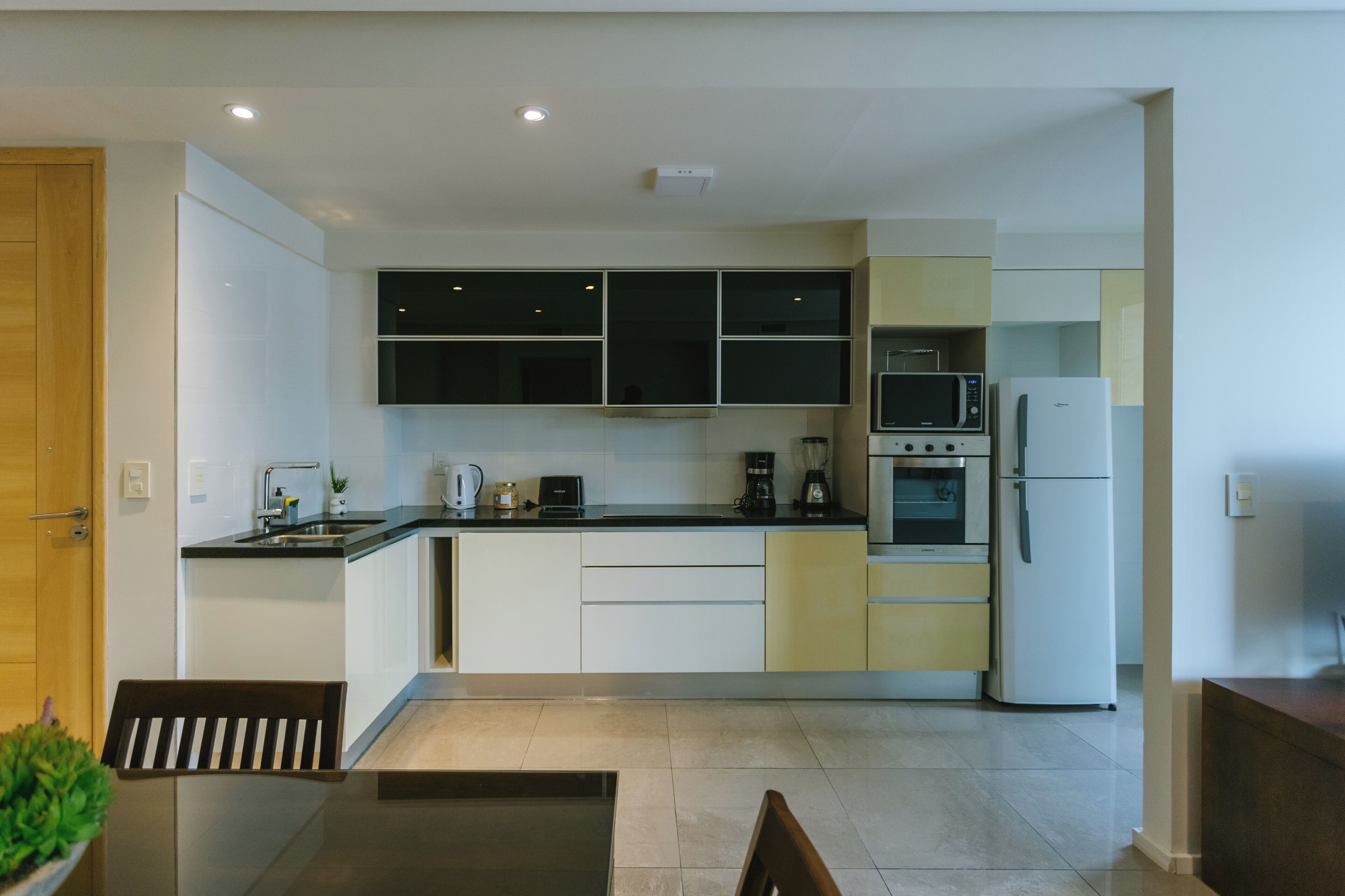 Een moderne keuken met witte en gele kastjes, een magnetron, oven en koelkast. Er staat een eettafel met stoelen en een potplant.