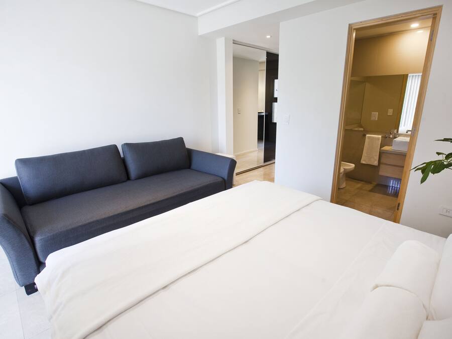 Une chambre à coucher moderne avec un lit, un canapé et une salle de bain visible à travers une porte ouverte.