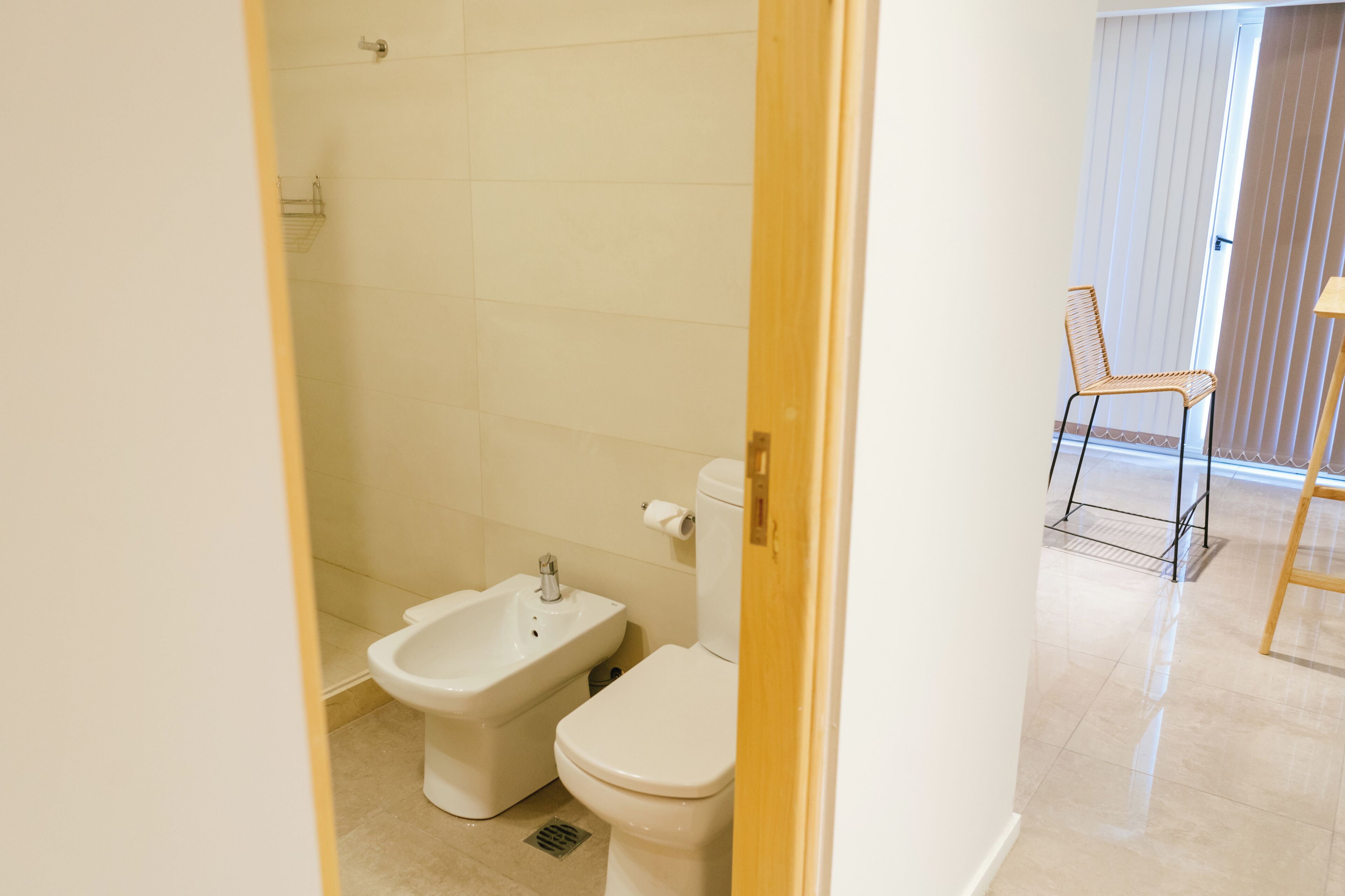 A bathroom with a toilet, bidet, and shower. There is a chair and a table in the adjacent room.