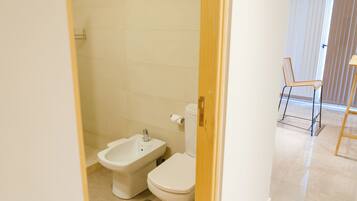A bathroom with a toilet, bidet, and shower. There is a chair and a table in the adjacent room.