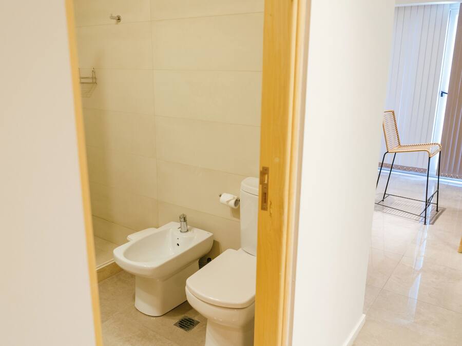 A bathroom with a toilet, bidet, and shower. There is a chair and a table in the adjacent room.