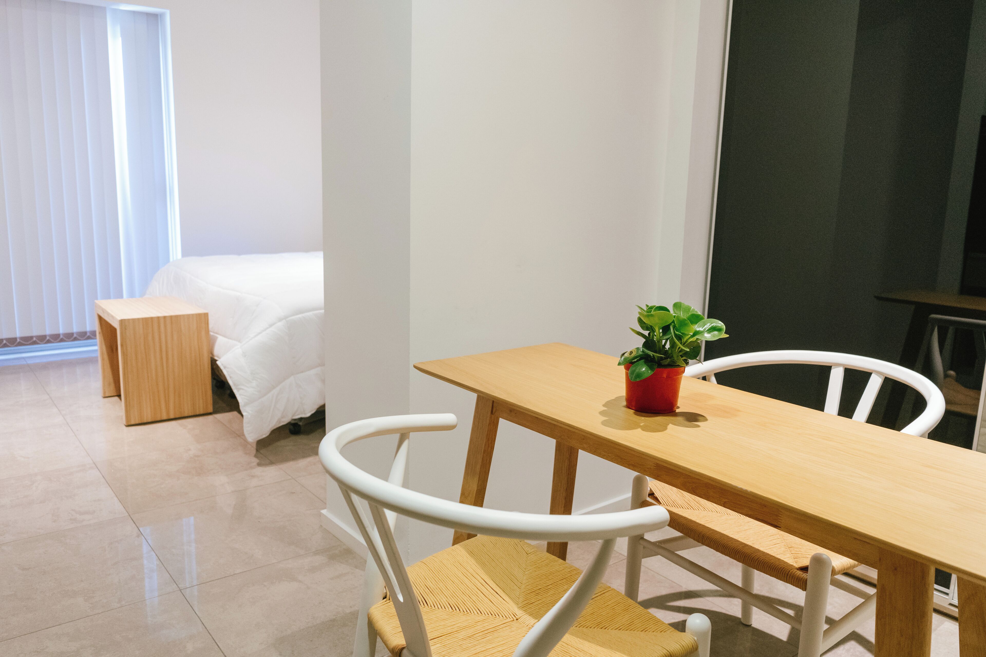 Una habitación de hotel moderna con una mesa de comedor de madera, sillas blancas, una pequeña planta en maceta, una cama y un ventanal con persianas.