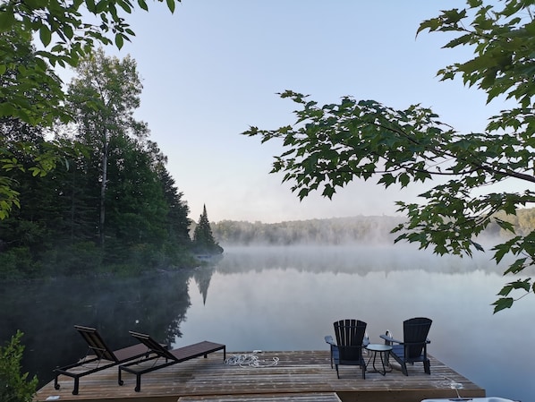 Une jetée en bois, agrémentée de chaises longues et d’une table, d’où l’on peut admirer un lac paisible, avec une forêt dense en arrière-plan.