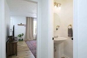 Un pasillo que da a un baño con lavamanos, espejo y argolla para toallas. En la habitación contigua, hay un televisor sobre un soporte.