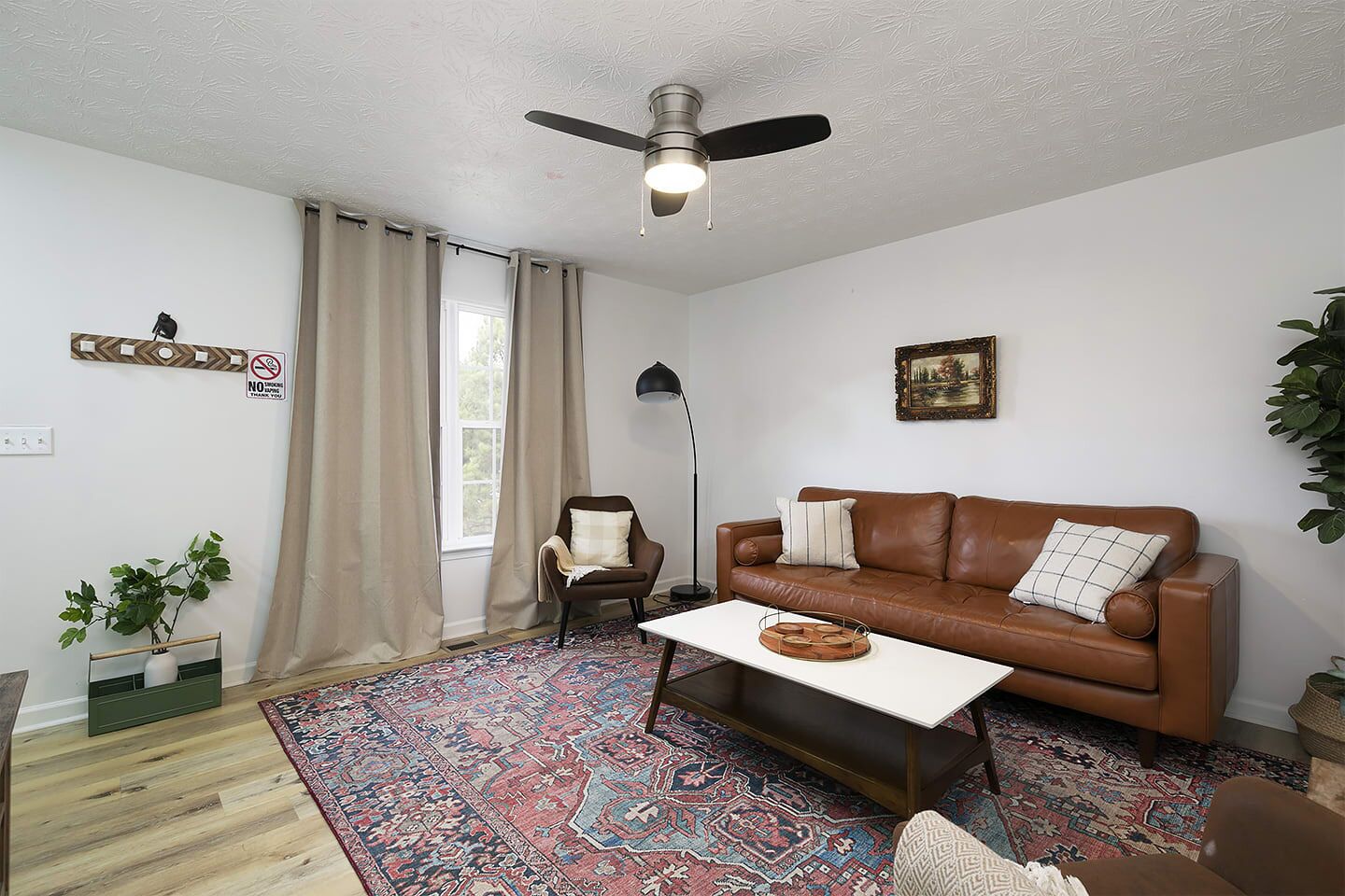 A living room with a brown leather sofa, a white coffee table, a patterned rug, a ceiling fan, and a window with beige curtains.