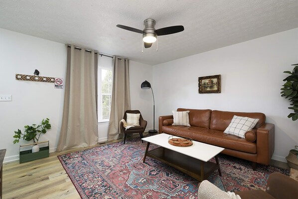 Una sala de estar con un sofá de cuero marrón, una mesa de centro blanca, un tapete estampado, un ventilador de techo y una ventana con cortinas beige.
