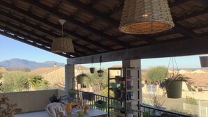 Un patio cubierto al aire libre con una mesa y sillas, rodeado de plantas en macetas y cestas colgantes.