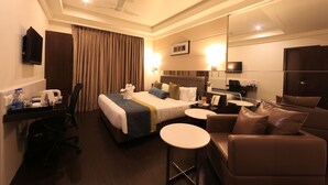A hotel room with a bed, a desk with a chair, a small round table, a sofa, a ceiling fan, and a large mirror. - Hotel Regalia Tirupati (Tirupati)
