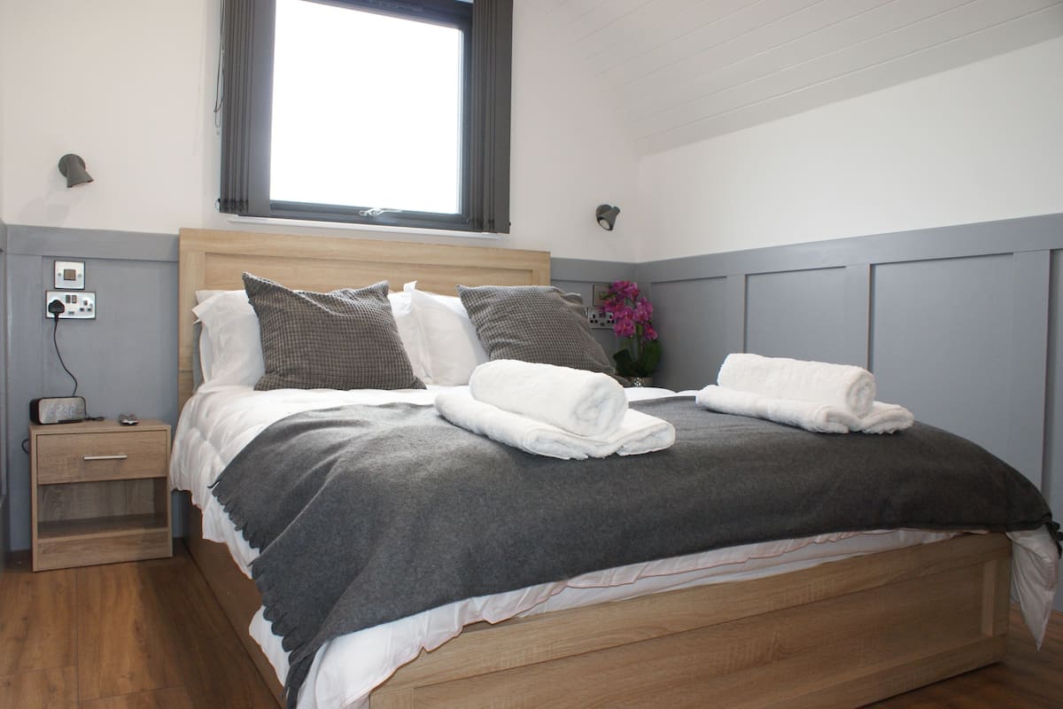 Una cama bien hecha con una manta y almohadas grises, una mesita de noche con una lámpara, y una ventana con cortinas.