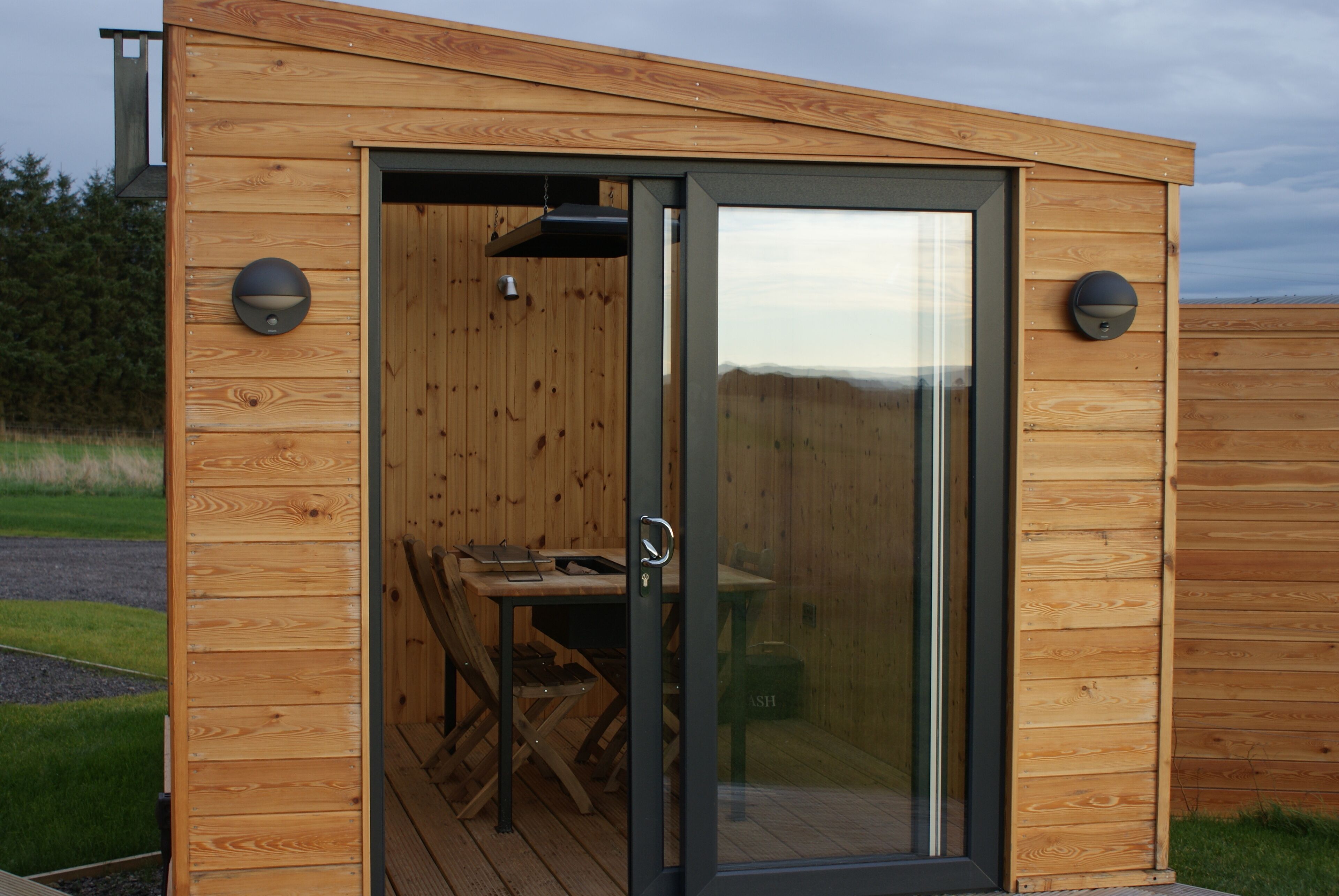 A wooden cabin with a sliding glass door, a table, and two wall-mounted lights.