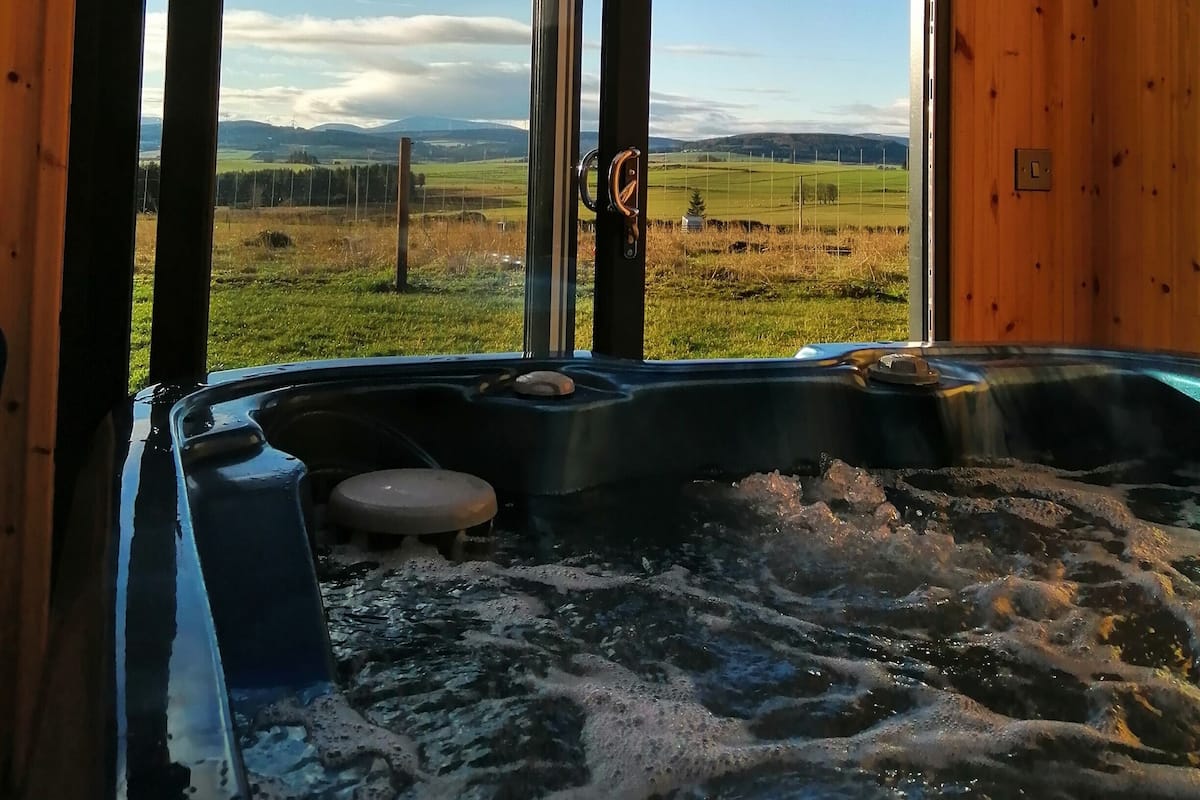 Una cabaña de madera con un jacuzzi y un ventanal grande que ofrece vistas a un campo de césped y montañas distantes.