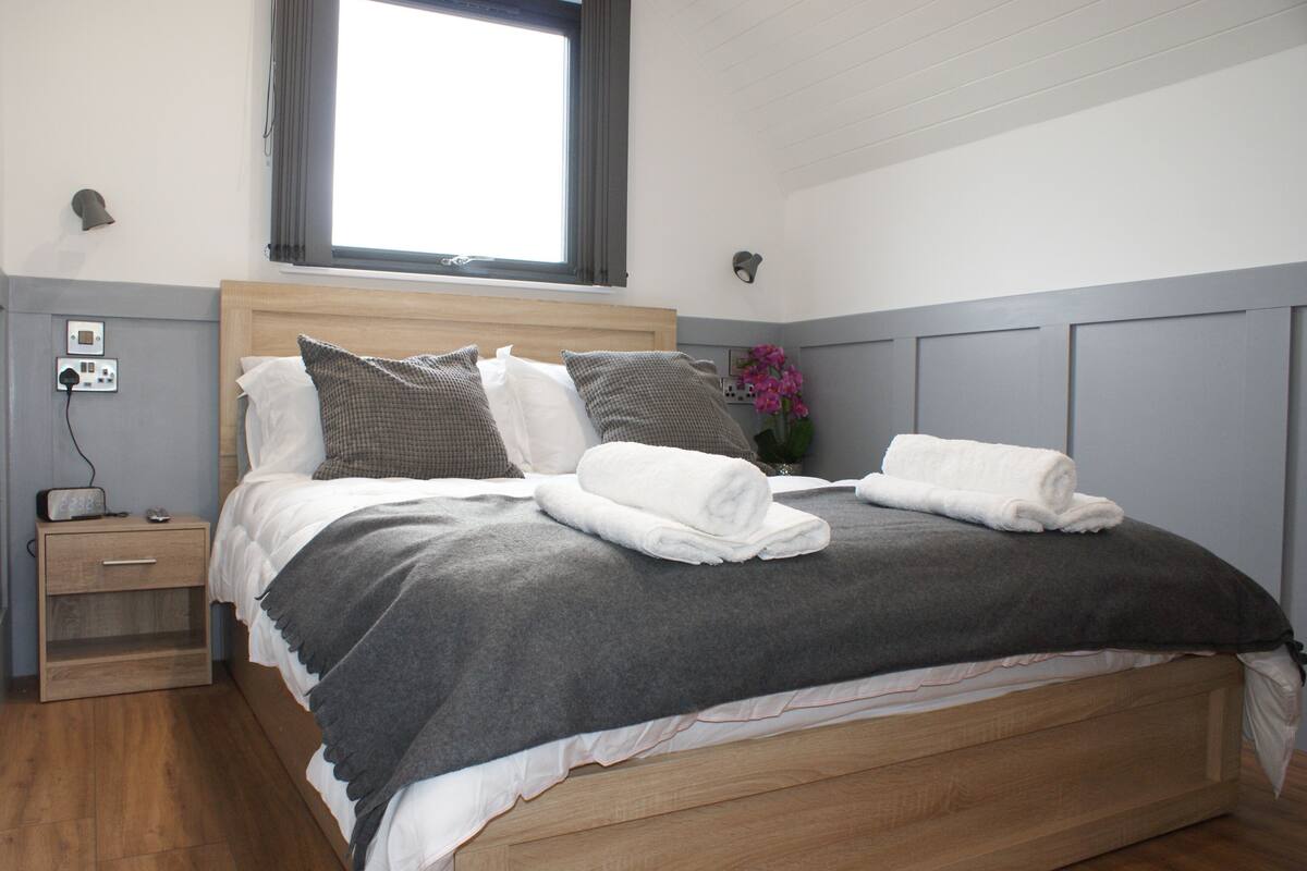 Una cama bien hecha con una manta y almohadas grises, una mesita de noche con una lámpara, y una ventana con cortinas.