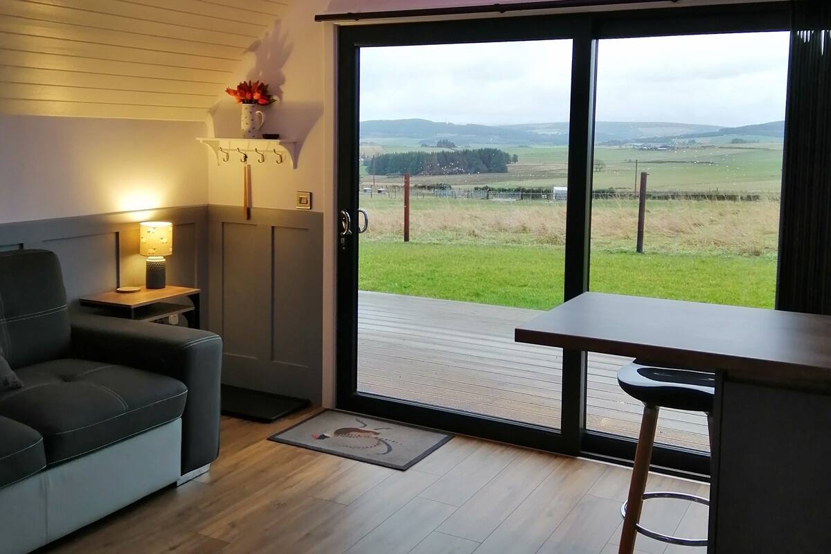 Una sala de estar moderna con un sofá, una mesa y vistas a un paisaje verde a través de un gran ventanal corredero.