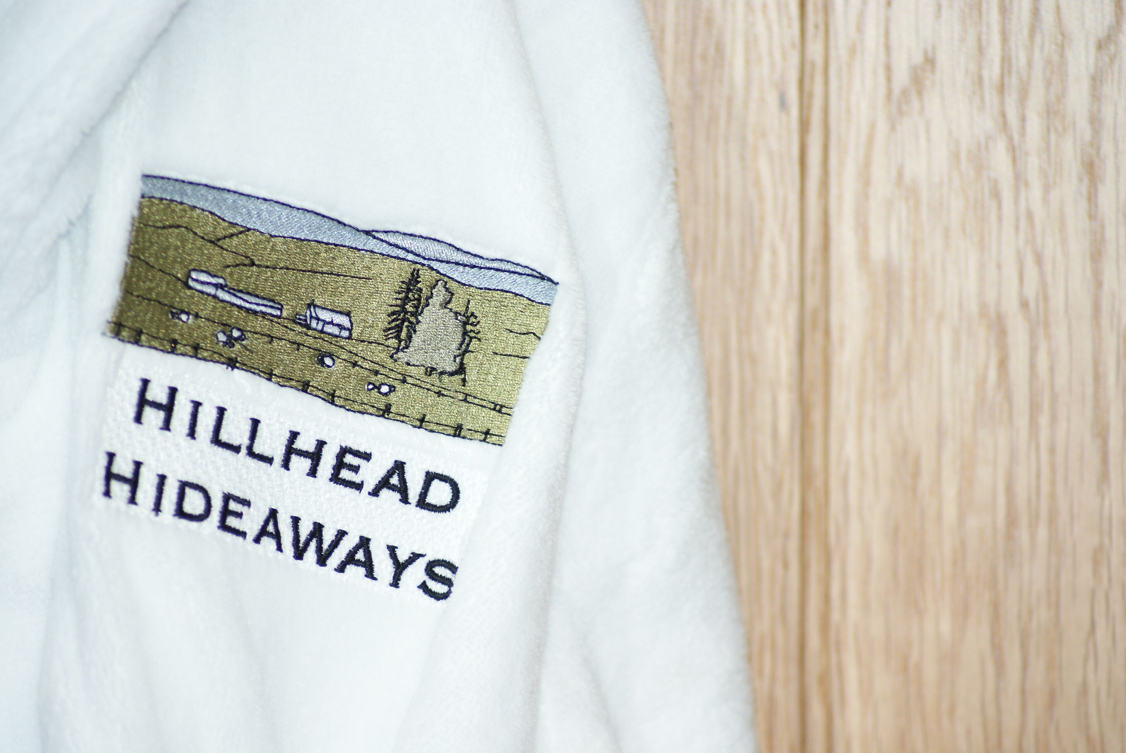 A white garment with embroidered text "HILLHEAD HIDEAWAYS" and a landscape illustration.