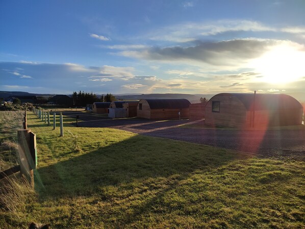 Una hilera de cabañas de madera con un camino de grava que las conduce, al pie de colinas bajo un cielo despejado.
