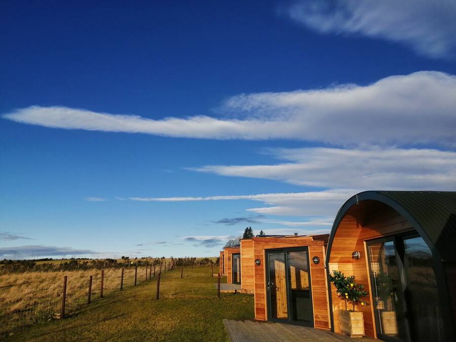 A modern wooden cabin with a large glass door and a curved roof, surrounded by a fenced field.