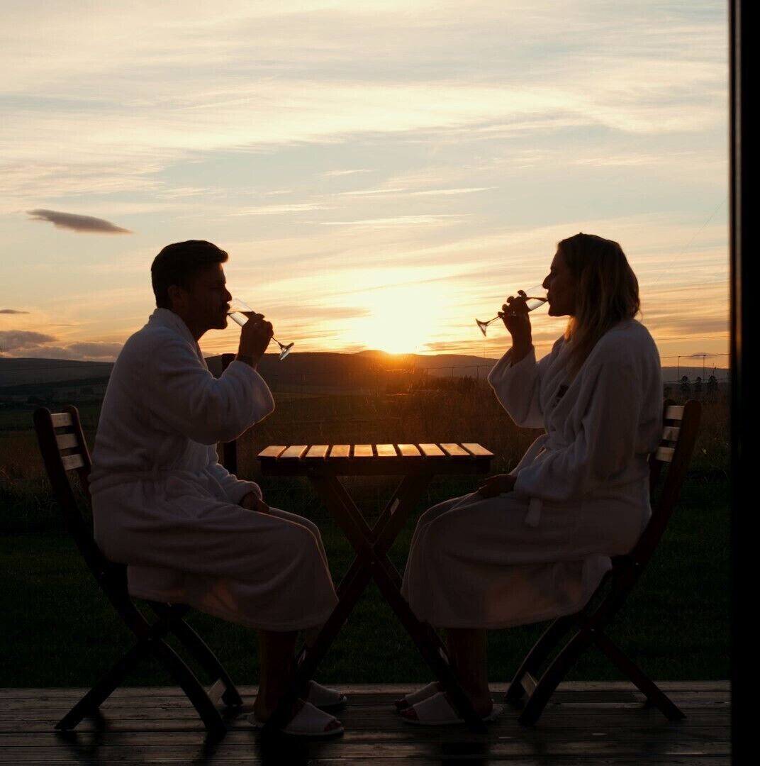 Dos personas vestidas de blanco sentadas en una mesa de madera al aire libre durante el atardecer.