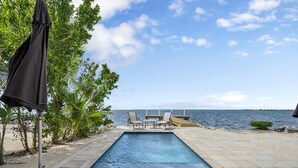Una piscina rectangular con vista al mar, rodeada por una terraza con baldosas, una sombrilla y sillas de descanso.