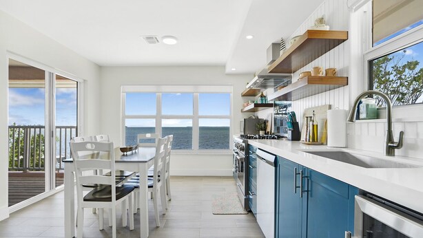 Una cocina moderna con vistas al mar, que cuenta con armarios blancos, un lavamanos de acero inoxidable y un comedor con sillas blancas.