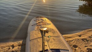Una tabla de paddleboard en una playa arenosa con una masa de agua tranquila y una puesta de sol al fondo.