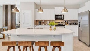 Una cocina moderna con armarios blancos, una isla central con taburetes de bar, iluminación colgante y electrodomésticos de acero inoxidable.