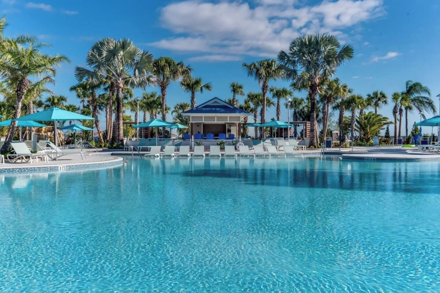 Piscina azul cristalina com espreguiçadeiras e guarda-sóis, rodeada por palmeiras e um edifício com telhado azul.
