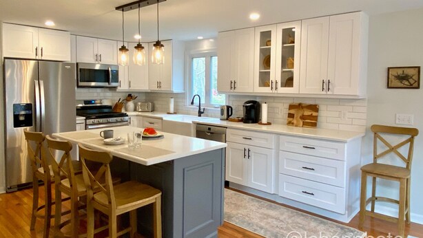 A modern kitchen with white cabinetry, a central island with bar stools, a stainless steel refrigerator, and a microwave. - Lovely three bedroom home located in desirable Bayside neighborhood! (South Dennis)