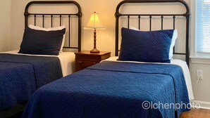 A bedroom with two beds, each with a dark blue quilted bedspread and matching pillows. There is a wooden nightstand with a lamp between the beds. - Lovely three bedroom home located in desirable Bayside neighborhood! (South Dennis)