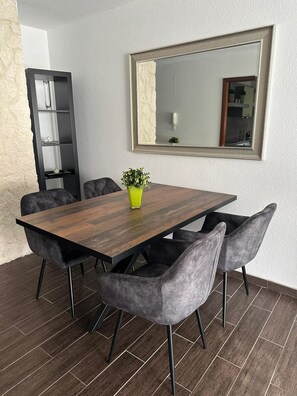 Ein Essbereich mit einem Holztisch, vier grau gepolsterten Stühlen, einer Zimmerpflanze und einem großen Spiegel an der Wand.