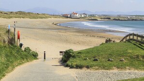 Une route côtière menant à une plage, avec un pont en bois, un sentier sablonneux et une zone herbeuse agrémentée d’un banc.