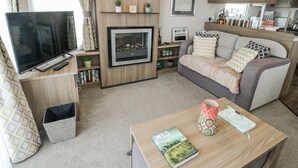 Un salon moderne avec un téléviseur à écran plat, une cheminée, un canapé, une table basse avec un livre et une tasse, et un coin cuisine à l’arrière-plan.