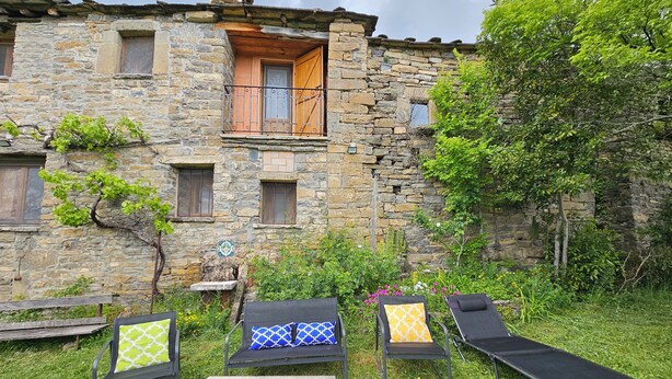 A stone building with a balcony, outdoor seating with cushions, and a garden area. - Comfortable house in the Pyrenees with views (Ainsa-Sobrarbe)