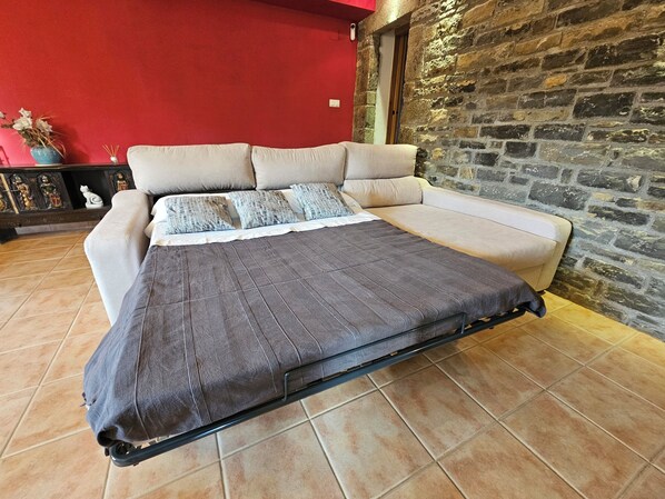 A bed with a grey mattress and a beige sofa against a red wall, with a stone wall on one side. - Comfortable house in the Pyrenees with views (Ainsa-Sobrarbe)