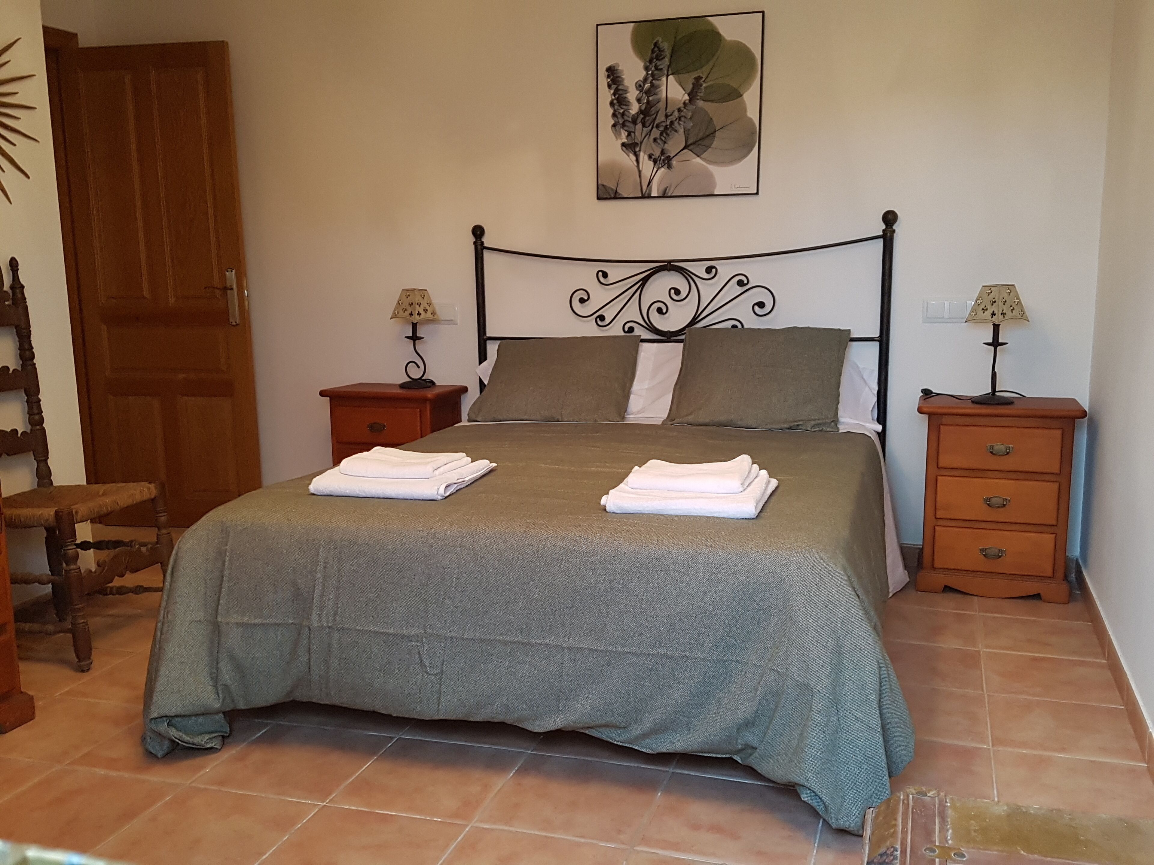 A neatly made bed with a grey bedspread and two folded white towels, a wooden nightstand with a lamp, a wooden chair, and a wooden door.