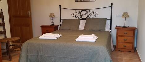 A neatly made bed with a grey bedspread and two folded white towels, a wooden nightstand with a lamp, a wooden chair, and a wooden door.