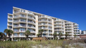 Un hôtel à plusieurs étages doté de balcons, situé près d’une plage de sable fin, où la végétation est clairsemée.