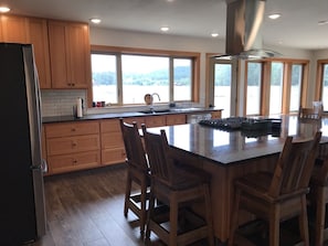 A modern kitchen with wooden cabinets, a large island with bar stools, a stainless steel range hood, and large windows offering a view of the outdoors.