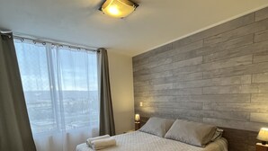 A modern hotel room with a large bed, a wooden headboard, a nightstand with a lamp, and a view of the city through the window.