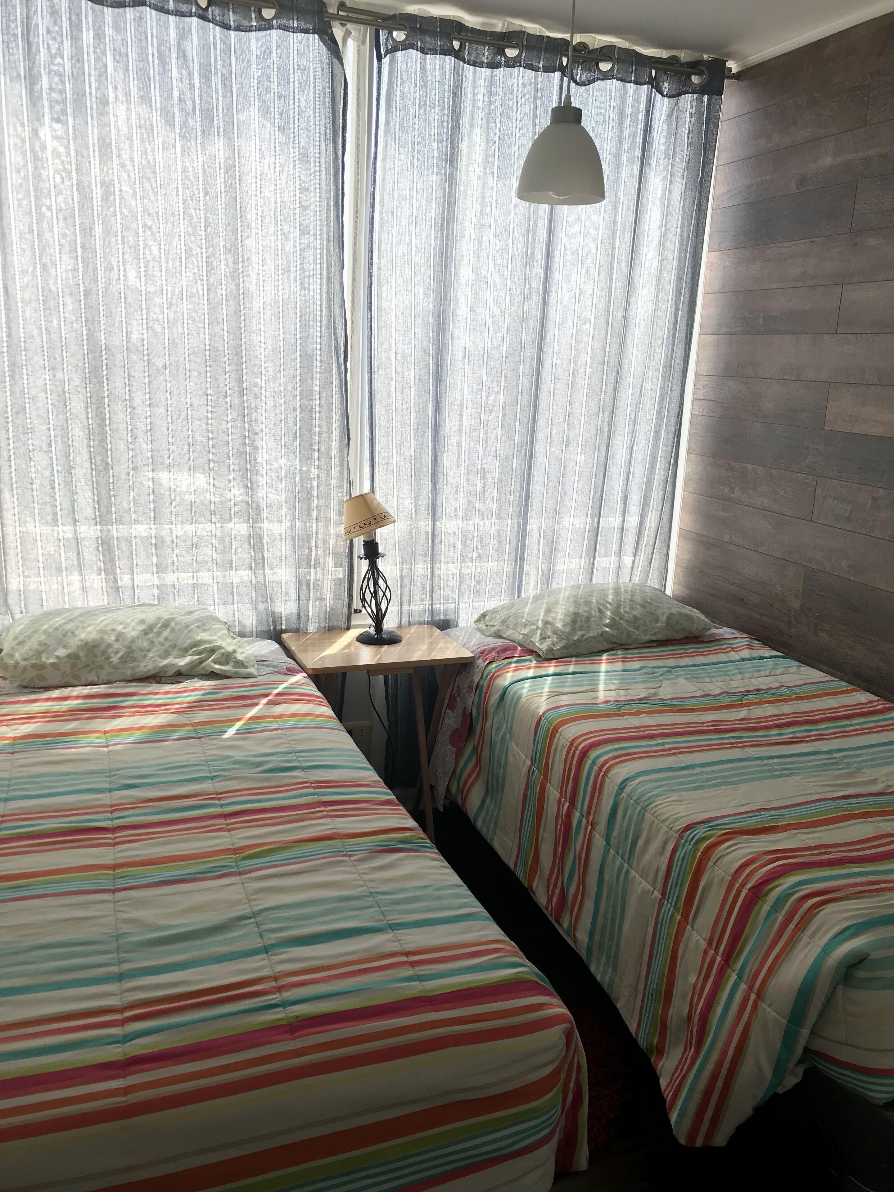 A room with two beds, a small table, a lamp, and sheer curtains.