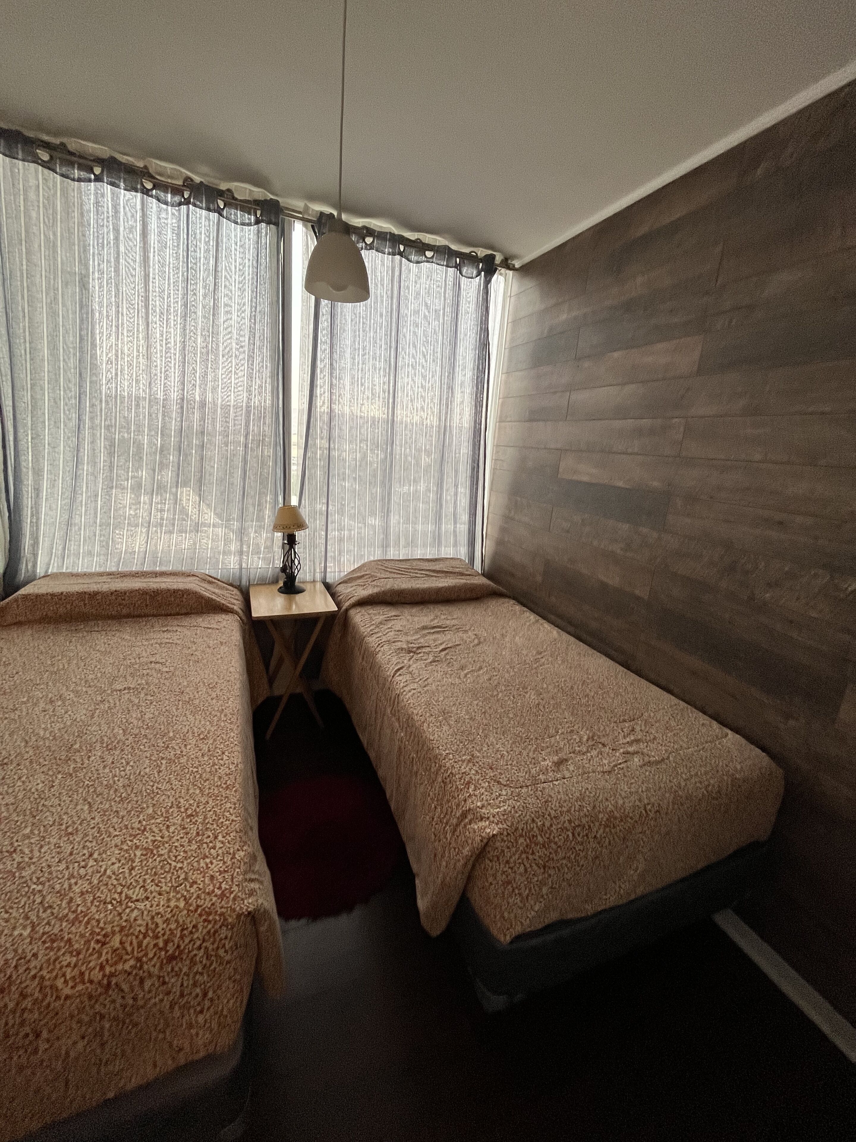 A room with two single beds, a small table with a lamp, and a window with sheer curtains.