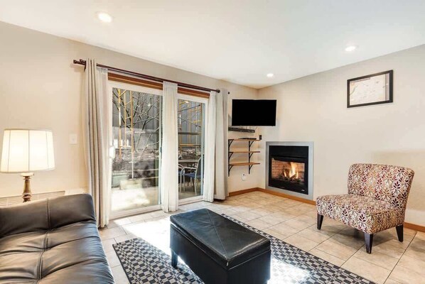 A living room with a sofa, a patterned armchair, a black ottoman, a fireplace, and a television mounted on the wall.