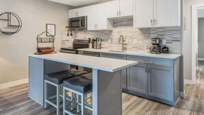 A modern kitchen with grey cabinets, a stainless steel microwave, and a built-in oven. There is a bar area with stools and a coffee machine.
