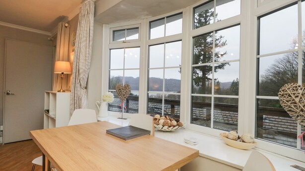 Un coin repas avec une table en bois, des chaises, une lampe, un vase de fleurs et une vue sur un paysage enneigé, visible à travers de grandes fenêtres.