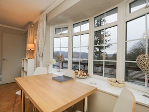 A dining area with a wooden table, chairs, a lamp, a vase with flowers, and a view of a snowy landscape through large windows. - Lake View (Windermere)