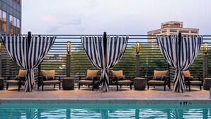 Un área de piscina en la azotea con cortinas a rayas y sillones de descanso, cielo despejado y un edificio al fondo.