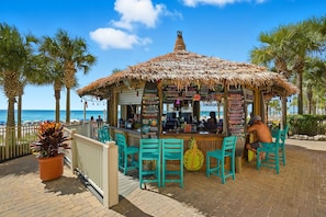 A beachside bar with a thatched roof, turquoise chairs, and a menu board.