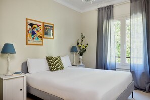 A neatly made bed with white linens, two pillows, a bedside table with a lamp, a framed poster on the wall, and a window with curtains. - Casa Cosi - Casanova 1 (Barcelona)